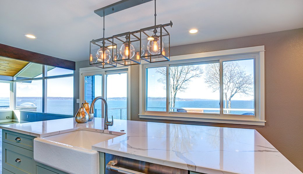 A boxy, modern light fixture with three lightbulbs illuminates a white marble topped kitchen island in a beige kitchen.